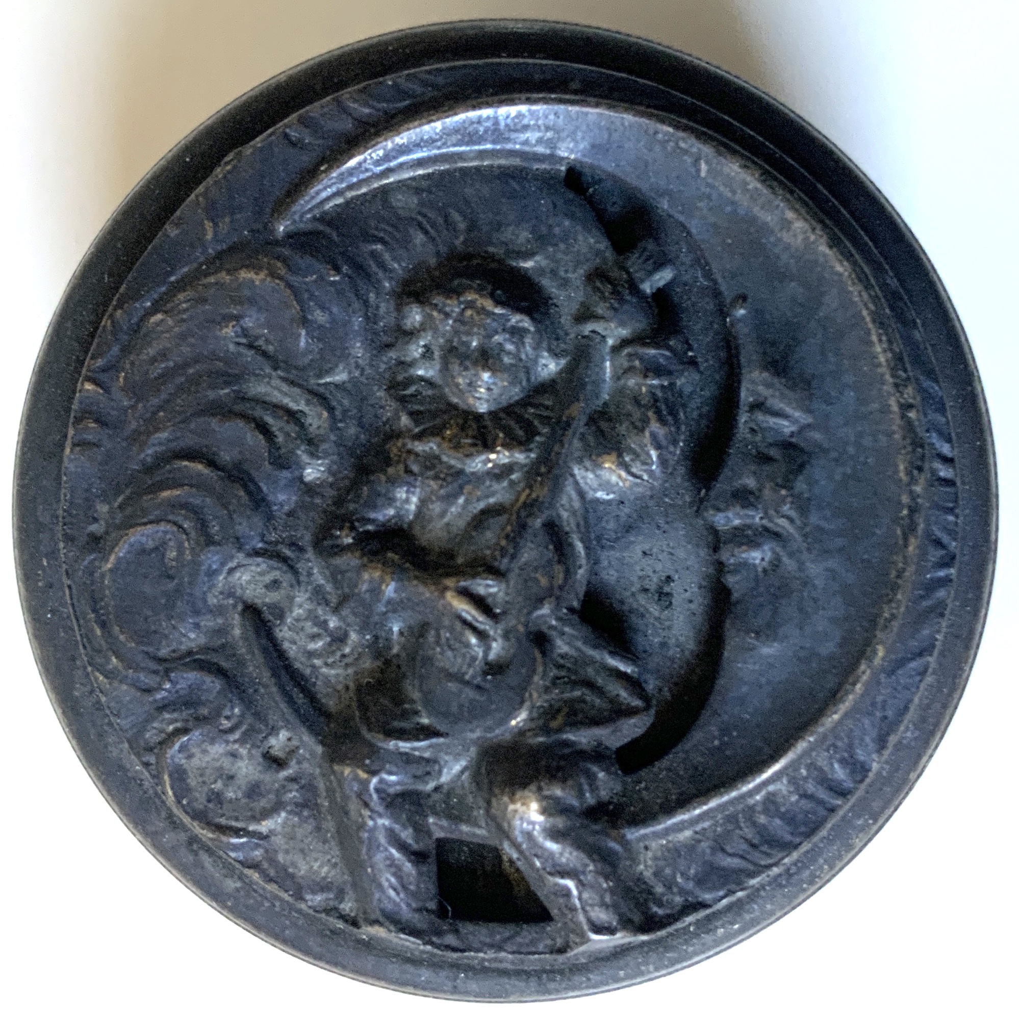 M290	AN EARLY SMALL PEWTER CONTAINER WITH A RELIEF ON TOP OF A CHERUB BOY SITTING IN THE LOWER HOOK OF A SMILING CRESCENT MOON PLAYING GUITAR